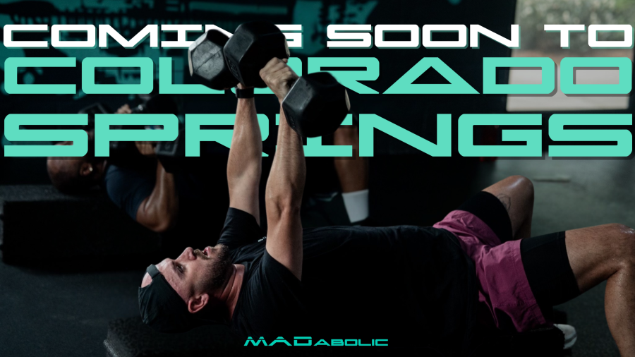 Athletic male performing dumbbell bench press. Text around him says "Coming soon to Colorado Springs" and the MADabolic wordmark is at the bottom of the image.
